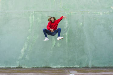 Portrait of young man in wearing red sweatshirt jumping in the air in front of green wall - AFVF04940