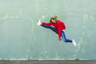 Porträt eines jungen Mannes in einem roten Sweatshirt, der vor einer grünen Wand in die Luft springt - AFVF04941