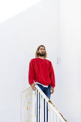 Portrait of bearded young man wearing red sweatshirt standing on stairs looking at distance - AFVF04945