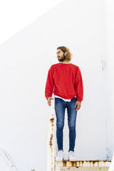 Portrait of bearded young man wearing red sweatshirt standing on rusty stairs - AFVF04946