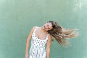 Blond young woman tossing her hair in front of green wall - AFVF04953