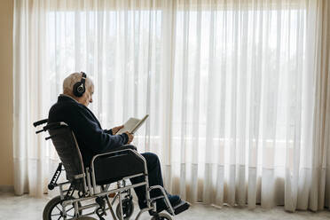 Älterer Mann sitzt im Rollstuhl und liest ein Buch, während er mit Kopfhörern Musik hört - JRFF03945