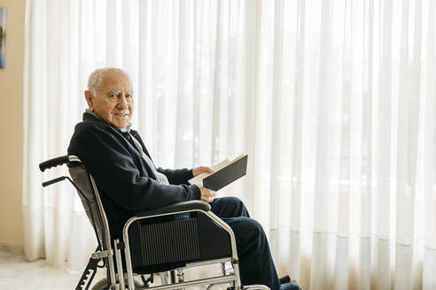 Porträt eines lächelnden älteren Mannes, der im Rollstuhl sitzt und ein Buch liest - JRFF03949