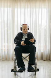 Porträt eines älteren Mannes, der im Rollstuhl sitzt und ein digitales Tablet benutzt, während er mit Kopfhörern Musik hört - JRFF03951