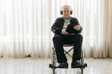 Porträt eines älteren Mannes im Rollstuhl sitzend mit digitalem Tablet, der mit Kopfhörern Musik hört - JRFF03952