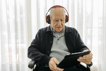 Älterer Mann sitzt im Rollstuhl und liest mit einem digitalen Tablet, während er mit Kopfhörern Musik hört - JRFF03953