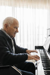 Senior man sitting in wheelchair playing piano at home - JRFF03961