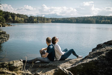 Happy couple sitting back to back at the lakeside, Forstsee, Carinthia, Austria - DAWF01079