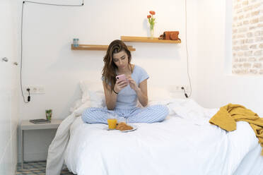 Junge Frau sitzt auf dem Bett und benutzt ihr Smartphone - ERRF02534