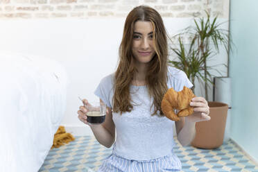 Junge Frau sitzt auf dem Boden und hält eine Tasse Kaffee und ein Croissant - ERRF02539