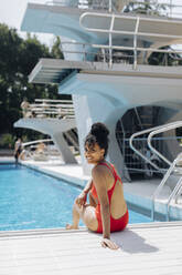 Portrait of smiling young woman wearing red swimsuit sitting at poolside - SODF00446