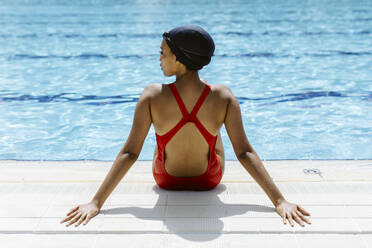 Rückenansicht einer jungen Frau in rotem Badeanzug und Badekappe, die sich nach dem Schwimmen am Beckenrand entspannt - SODF00459