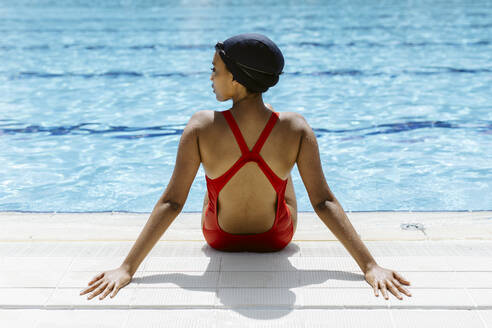 Rückenansicht einer jungen Frau in rotem Badeanzug und Badekappe, die sich nach dem Schwimmen am Beckenrand entspannt - SODF00459
