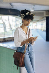 Porträt einer jungen Frau mit Ohrstöpseln, die in einer Passage auf ihr Mobiltelefon schaut - SODF00481