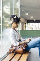 Porträt einer jungen Frau mit Ohrstöpseln, die auf einer Bank sitzt und auf ihr Mobiltelefon schaut - SODF00482