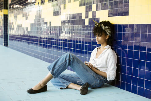 Portrait of young woman sitting on tiled floor listening music with earphones and smartphone - SODF00484