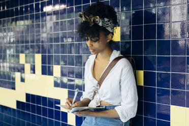 Junge Frau lehnt an einer gekachelten Wand und schreibt in ein Notizbuch - SODF00485