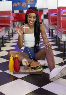 Junge Frau mit geflochtener Frisur sitzt mit ihrem Hamburger-Teller auf dem Boden und isst einen Chip - VEGF01275