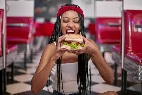 Young woman with braided hairstyle sitting on the floor and grabbing a big hamburger with her two hands - VEGF01278