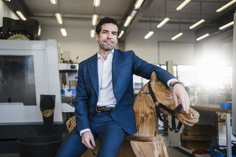 Porträt eines Geschäftsmannes auf einem hölzernen Schaukelpferd in einer Fabrik, lizenzfreies Stockfoto