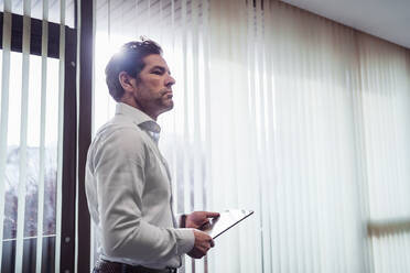 Businessman holding tablet at the window in office - DIGF09125