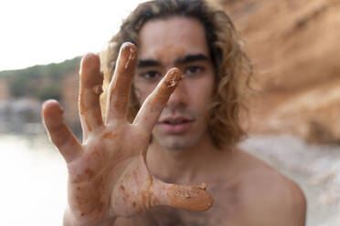 Young man showing his hand with mud on a beach - AFVF04983