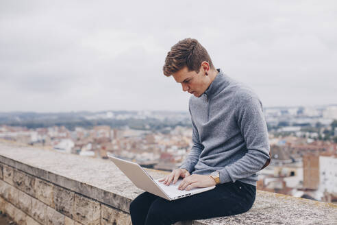 Blonder junger Mann sitzt an einer Wand und tippt auf seinem Laptop in Lleida - ACPF00652