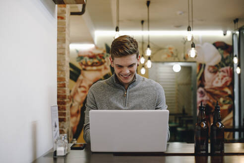 Junger Mann sitzt in einem Restaurant und tippt lächelnd auf seinem Laptop - ACPF00653