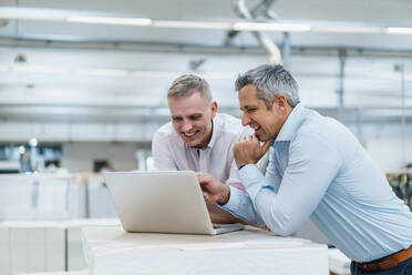 Zwei lächelnde Kollegen benutzen einen Laptop und diskutieren in einer Fabrik - DIGF09256