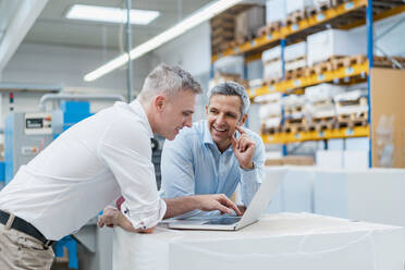 Two smiling colleagues using laptop and discussing in a factory - DIGF09258