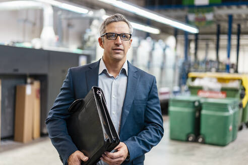 Porträt eines Geschäftsmannes mit Brille und Aktentasche in einer Fabrik - DIGF09283
