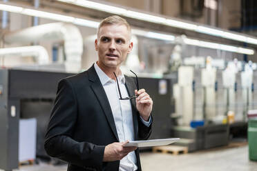 Businessman holding glasses and tablet in a factory looking around - DIGF09287