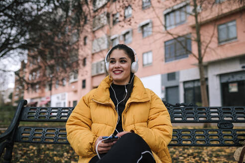 Porträt einer glücklichen jungen Frau, die im Herbst auf einer Bank sitzt und mit Kopfhörern und Smartphone Musik hört - GRCF00015