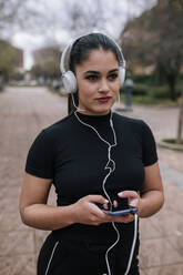 Porträt einer jungen Frau in schwarzer Sportkleidung, die mit Kopfhörern und Smartphone Musik hört - GRCF00016