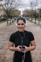 Portrait of happy young woman in black sportswear listening music with headphones and smartphone - GRCF00017