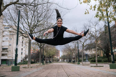 Portrait of young woman in black sportswear jumping in the air while listening music with headphones - GRCF00018