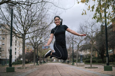 Porträt einer jungen Frau in schwarzer Sportkleidung, die in die Luft springt, während sie mit Kopfhörern Musik hört - GRCF00020