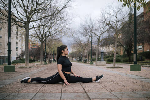 Junge Frau in schwarzer Sportkleidung macht einen Spagat auf dem Bürgersteig - GRCF00022