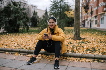 Portrait of smiling young woman listening music with headphones and smartphone in autumn - GRCF00023