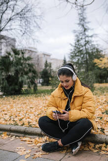 Junge Frau hört Musik mit Kopfhörern und Smartphone im Herbst - GRCF00024