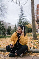 Porträt einer lächelnden jungen Frau, die mit Kopfhörern und Smartphone im Herbst Musik hört - GRCF00025