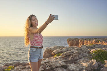 Young woman standing on cliff at the sea, using smartphone, taking selfie - EPF00648