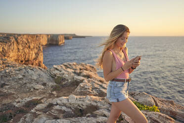 Young woman standing on cliff at the sea, using smartphone - EPF00650