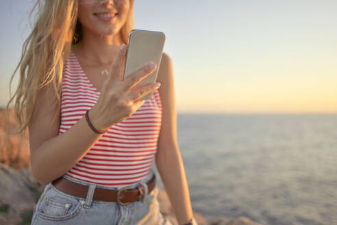 Junge Frau steht auf einer Klippe am Meer und benutzt ein Smartphone - EPF00651
