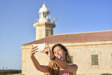 Young woman taking pictures of Punta Nati lighthouse, Menorca, Spain - EPF00658