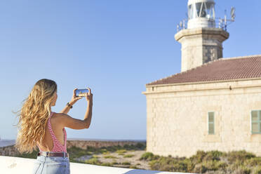 Young woman taking pictures of Punta Nati lighthouse, Menorca, Spain - EPF00659
