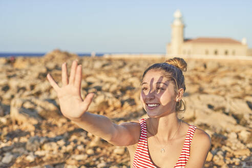 Junge Frau an einem felsigen Strand, die ihr Gesicht mit der Hand abschirmt, Menorca, Spanien - EPF00667