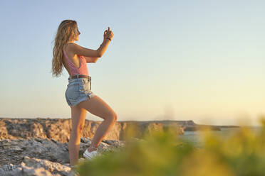 Junge Frau steht auf einer Klippe am Meer und benutzt ein Smartphone - EPF00672