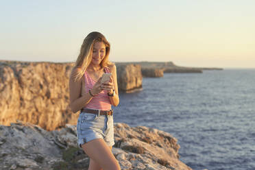 Young woman standing on cliff at the sea, using smartphone - EPF00673