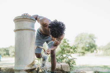 Young man during workout, drinking from fountain in a park - FBAF01237